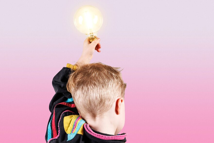The back of a young boy's head as he holds up an illuminated lightbulb in a pink room