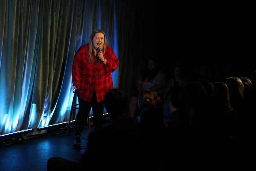 Stand-up comedian Mish Wittrup performing on a small stage in front of an audience seated in the dark.
