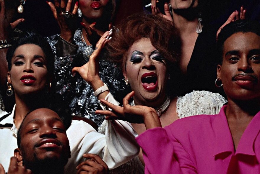 A still from the 1990 documentary Paris Is Burning, showing a crowd of people, several of them wearing full drag makeup and wigs.