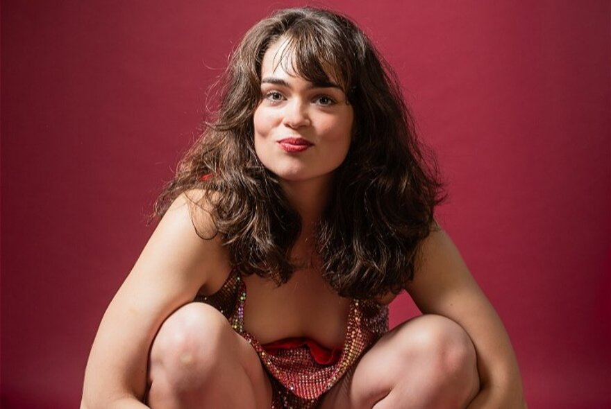 A woman crouching, wearing a gold sequined top and her long brown hair down around her shoulders; in front of a dark red background.