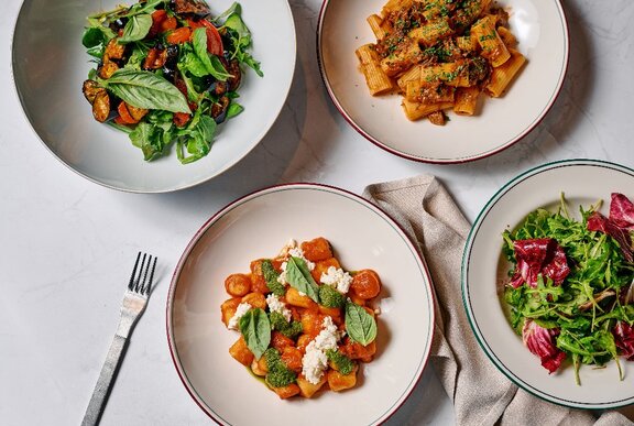 Four round plates on a marble table, one with a roast pumpkin salad, one with a rigatoni bolognese, one with a tomato gnocchi and one with a radicchio salad.
