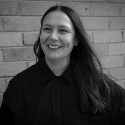 A smiling woman with long dark hair and wearing a dark top stands against a brick wall backdrop.