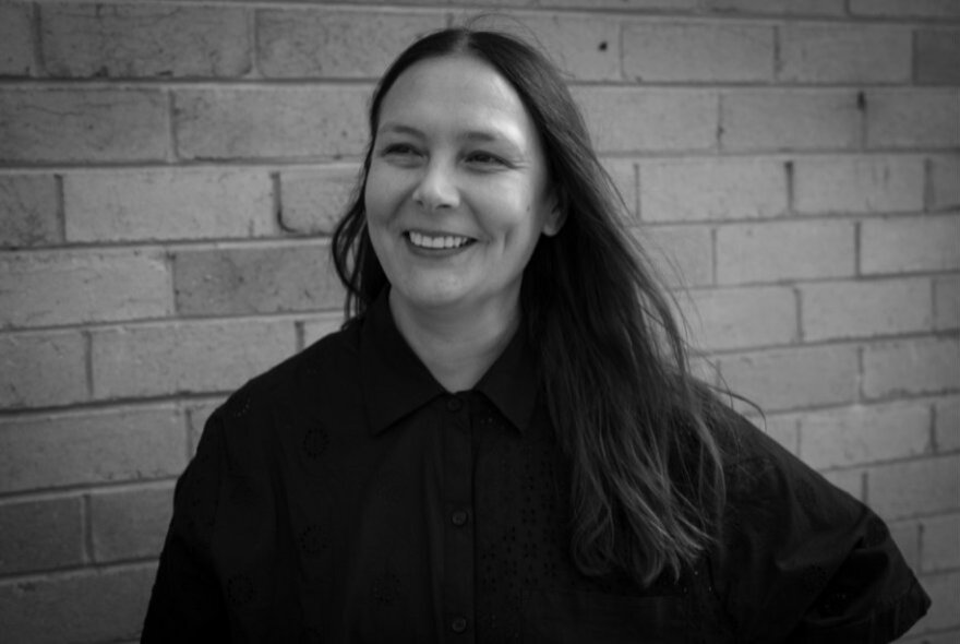 A smiling woman with long dark hair and wearing a dark top stands against a brick wall backdrop.