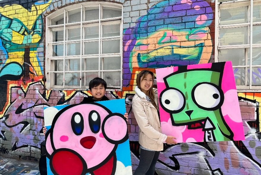 Two young people holding large cartoonish paintings in a laneway with street art on the walls.