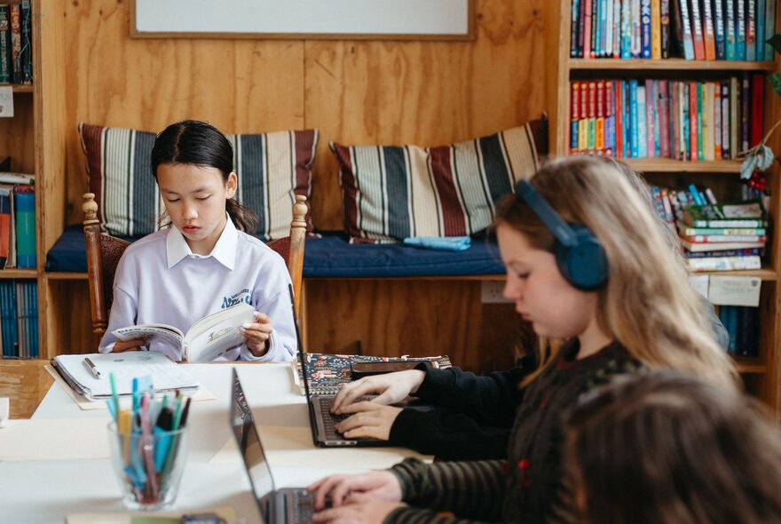 Children seated at a large table, one reading a book, the others on laptops.