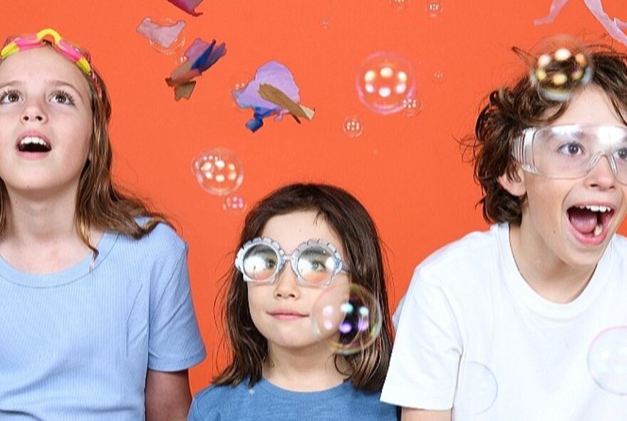 Three children, two with goggles on, looking at bubbles in awe, in front of a red wall.