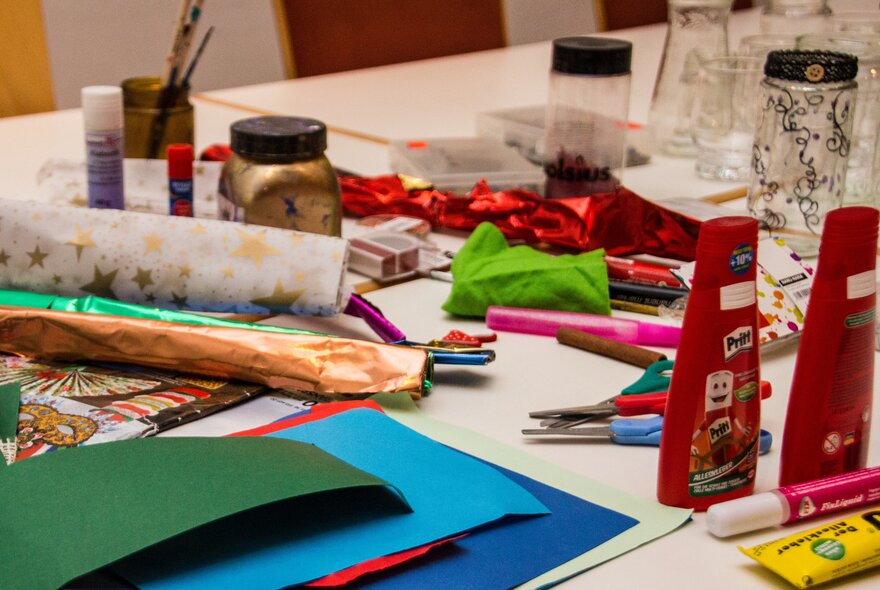 A table full of craft materials such as coloured card, paint, brushes, glue and printed paper. 