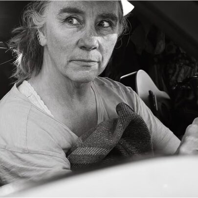 A woman seated at the wheel of a car, looking to the side with an alarmed expression.