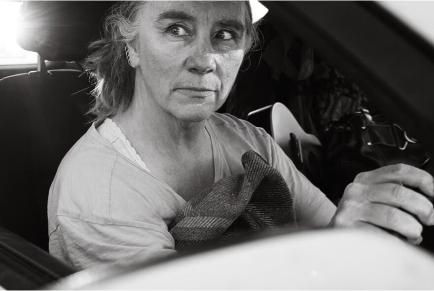 A woman seated at the wheel of a car, looking to the side with an alarmed expression.