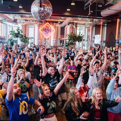 A large, energetic crowd of sports fans celebrating inside a crowded bar, taking part in a watch party for an NFL game.