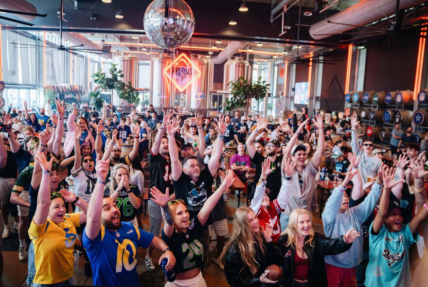 A large, energetic crowd of sports fans celebrating inside a crowded bar, taking part in a watch party for an NFL game.