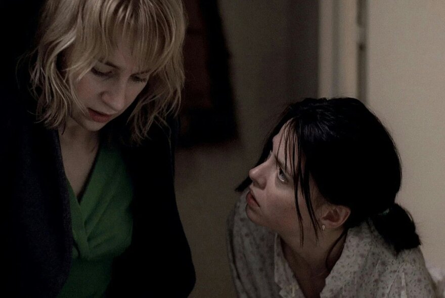 A still image from a film showing a woman looking up intently at another woman who is looking straight downwards. 