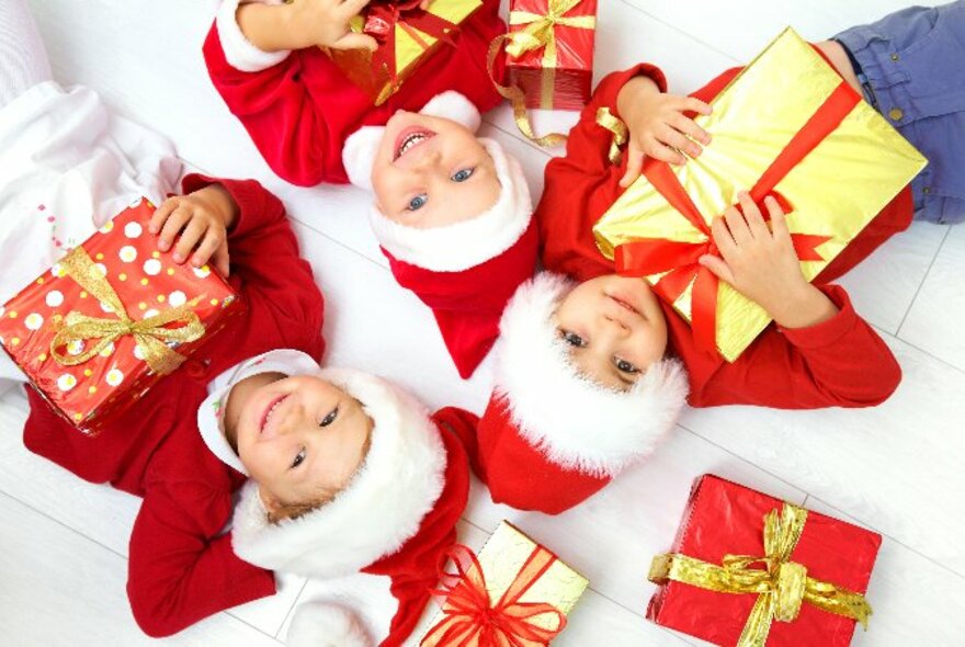 Overhead view of three young children, all wearing red tops and red Santa hats, lying on their backs looking up at the camera and holding wrapped parcels and presents to their chest.