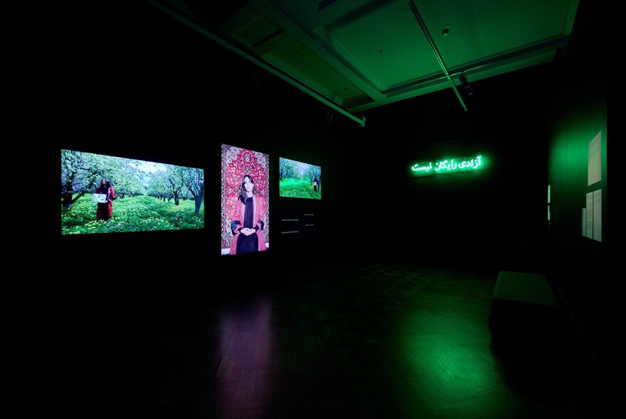 A darkened gallery space with three brightly coloured digital artworks on display.