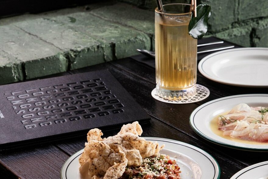 Plates of bar snacks and a cocktail in a long glass on a table, with a menu alongside.