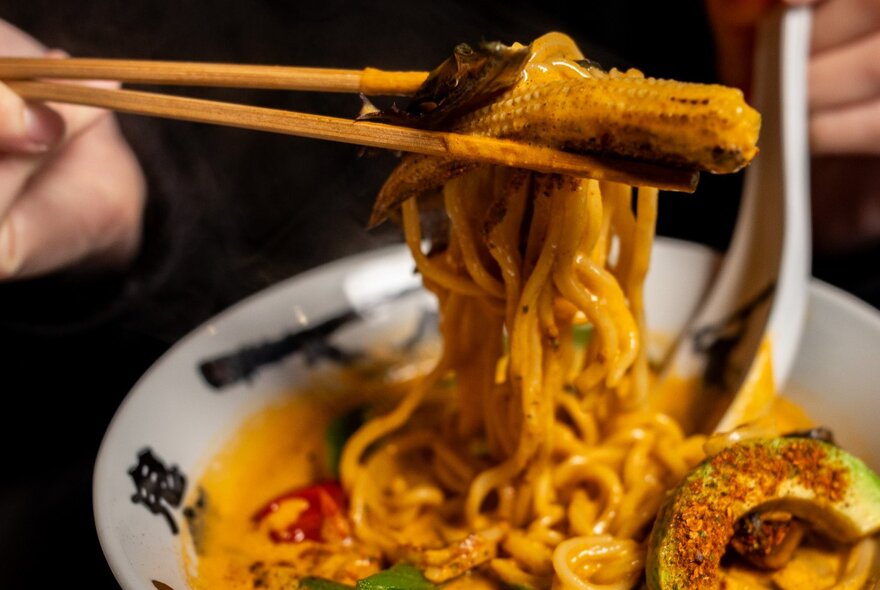 A pair of chopsticks holding noodles and a mini corn above a bowl of hot ramen.