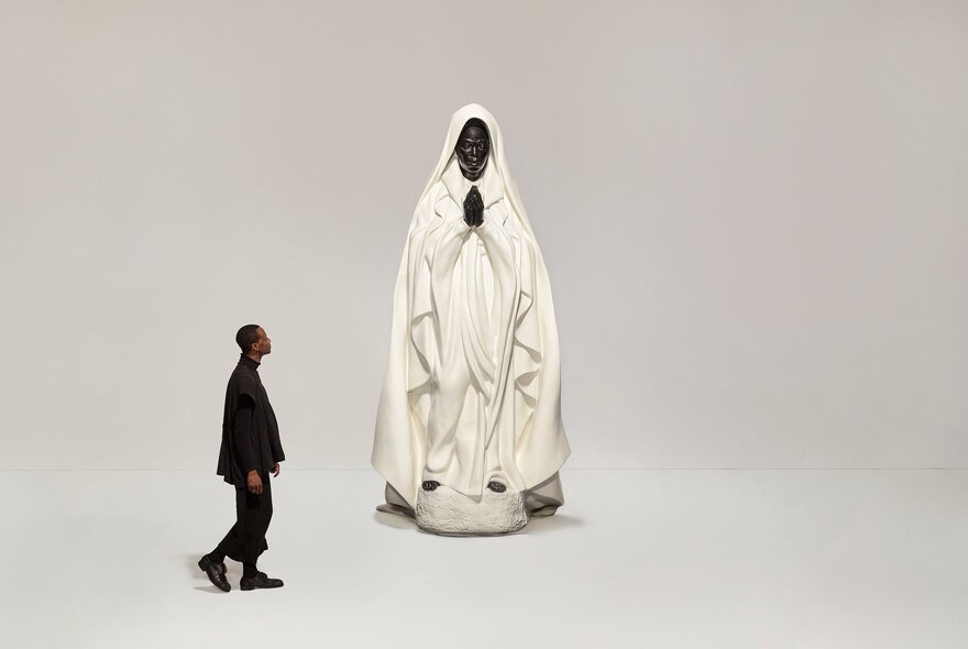 A large statue of a person in a white robe with their hands together in prayer and a man looking at the statue while walking by. 