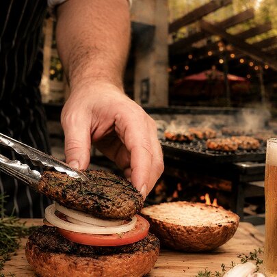 A commercial kitchen with hands and tongs placing a meat patty on top of onions in a burger bun, a glass of beer also visible. 