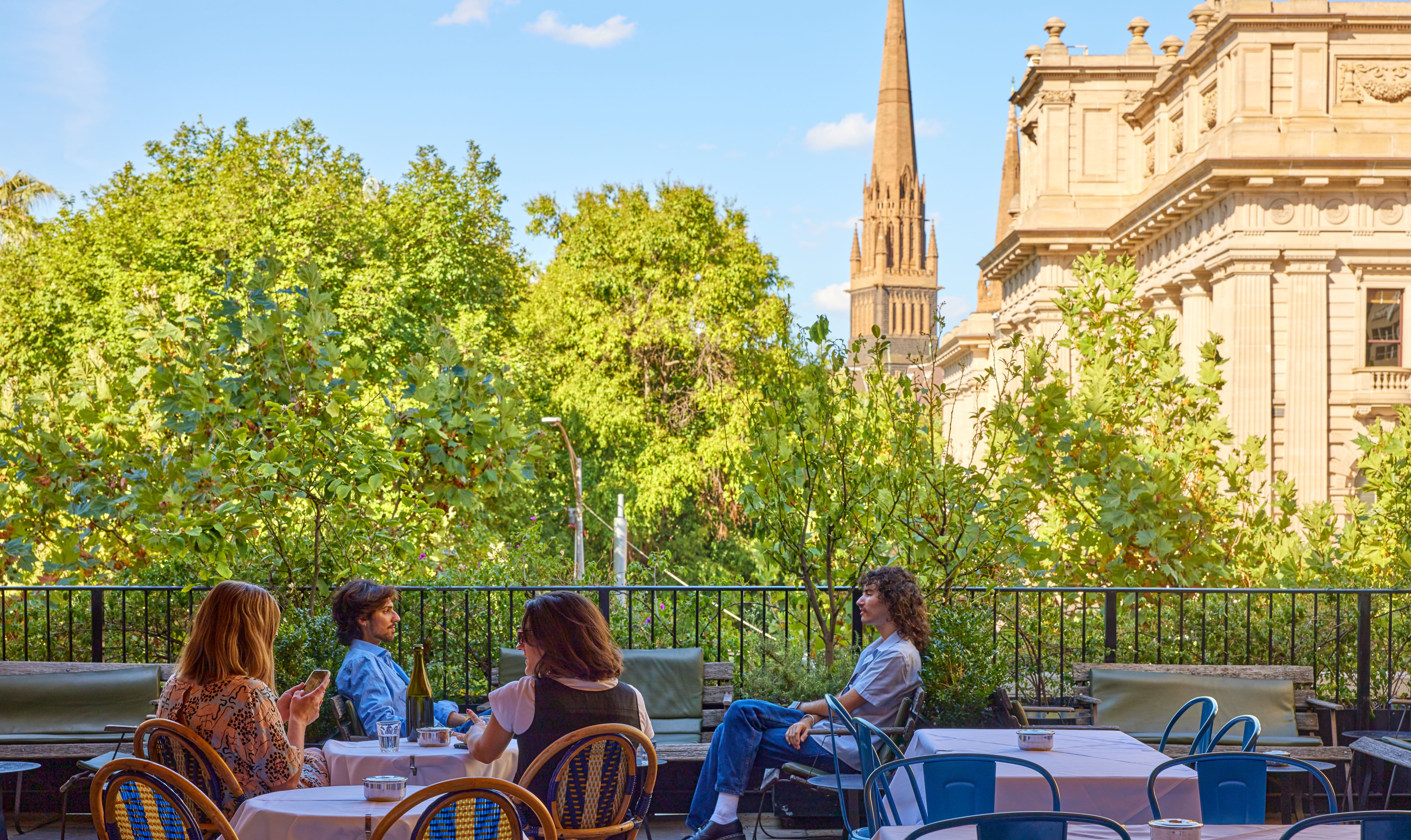 Four people sitting at a rooftop bar with a view of trees, a cathedral and a heritage building.