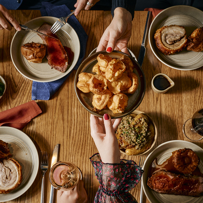 Someone passing a plate of Yorkshire puddings over a table set with rolled pork roast, veggies, wine and gravy.