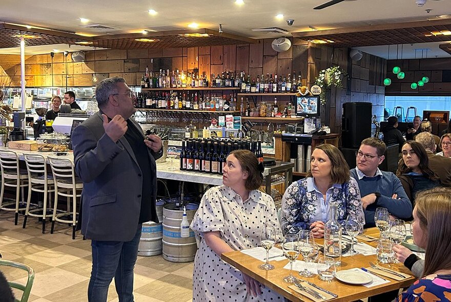 A person talking at a food and wine event inside a restaurant, while a table of diners face towards the speaker.