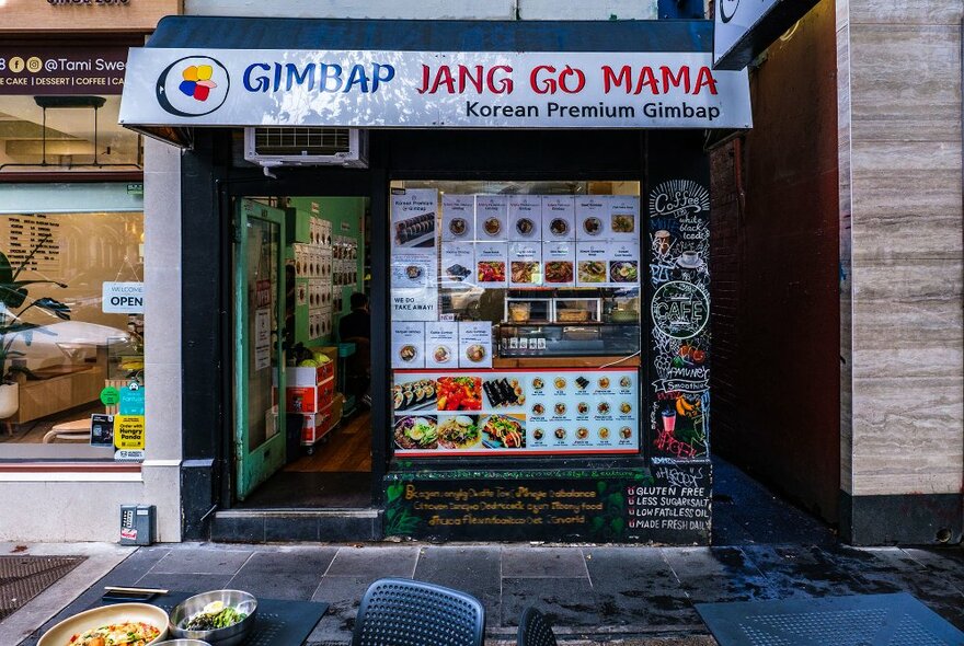 The exterior of a small city cafe with pictures of the menu on the windows and a table with food outside.