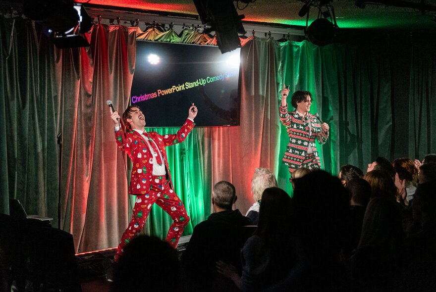 Two people on stage, wearing festive-themed suits outfits, performing to a seated audience in a small theatre room in front of red and green coloured curtains.