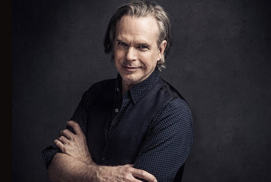 Singer Rick Price smiling and leaning forward slightly with his arms folded against a dark background. 