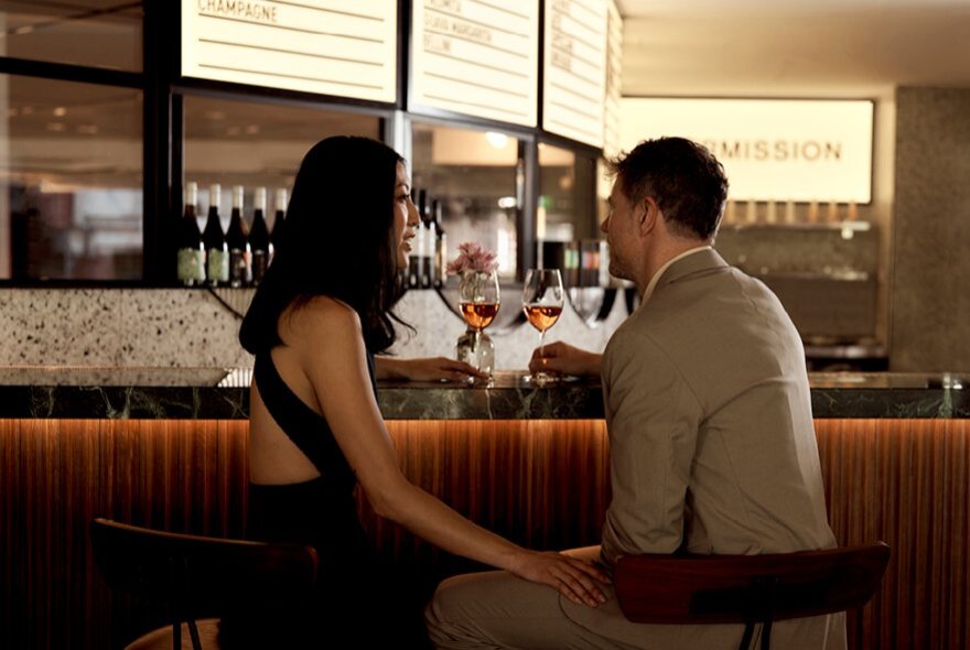 A young couple seated at a bar that looks like a cinema foyer, each holding a wine glass.