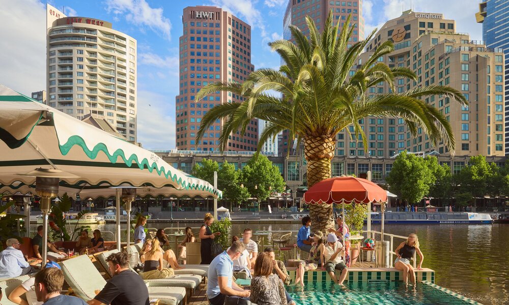 A group of people are sitting on sun loungers and by the pool on a floating bar on a river. There is a palm tree in the background as well as the city skyline. 