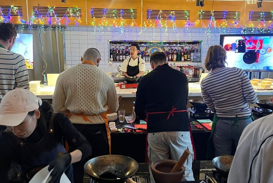 The back of people participating in a cooking class with a chef standing up front. 