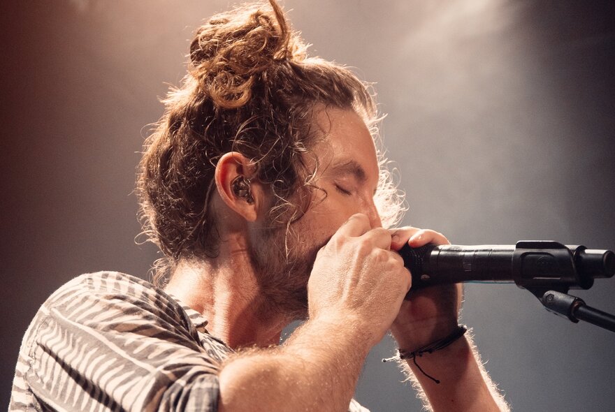 Profile of the singer Jeremy Loops, his hair is piled on top of his head in a loose bun, his eyes are closed and he is playing a harmonica which he holds close to the microphone stand.