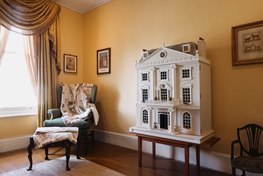 Georgian-style doll's house on a stand in a room with period furnishings including armchair and footstool, draped curtains and paintings.