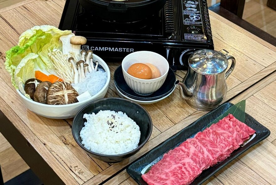 A plate of raw sliced beef waiting to be cooked in a hot pot on a portable cooktop resting on a restaurant table, with side dishes of rice, egg and raw vegetables.