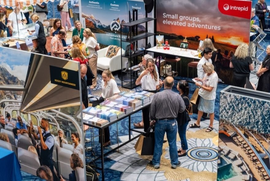 Stallholders and expo visitors browsing a stand beside travel posters and advertisements.