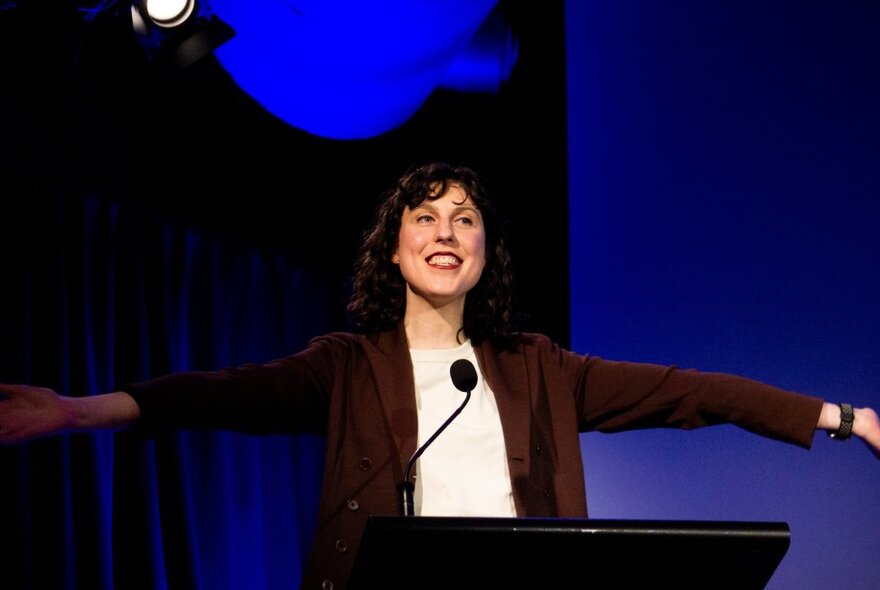 A woman standing behind a speaker's podium with a microphone, arms outstretched and smiling.