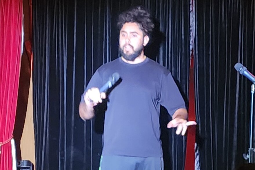 A male stand-up comedian holding a microphone and performing on a stage.