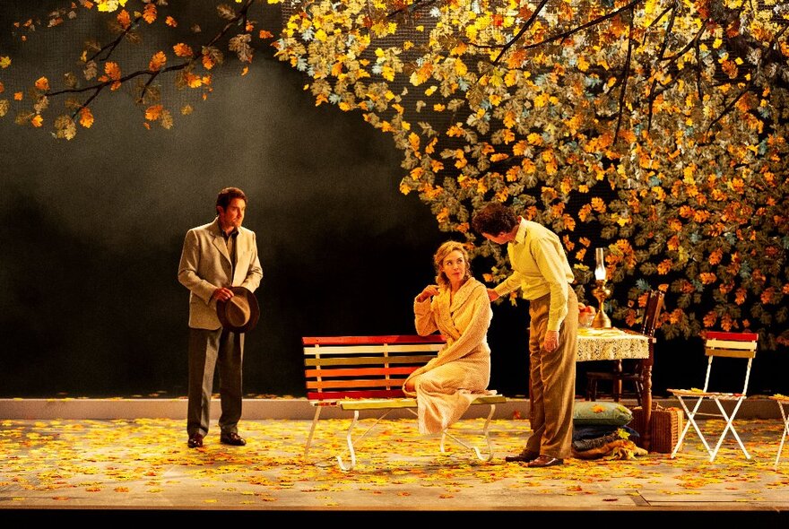 Three actors on stage in 1940s-style costumes with a set design depicting an autumn picnic.