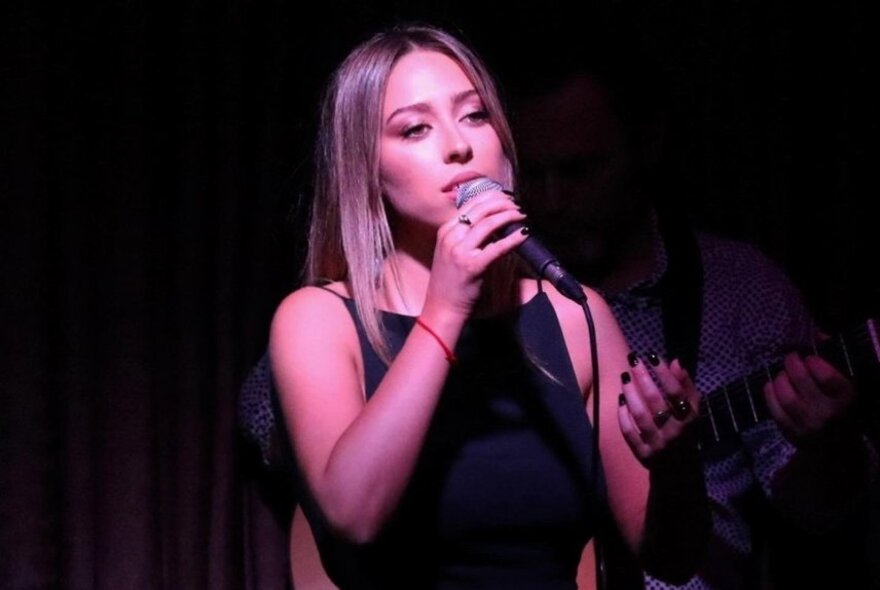 Performer Jemma Cher singing on stage under pink lights, holding a microphone.