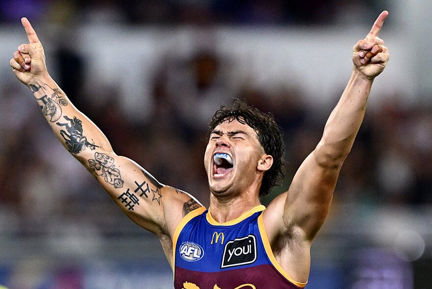 A Brisbane Lions AFL player celebrating during a match with tattooed arms stretched into the air, yelling.
