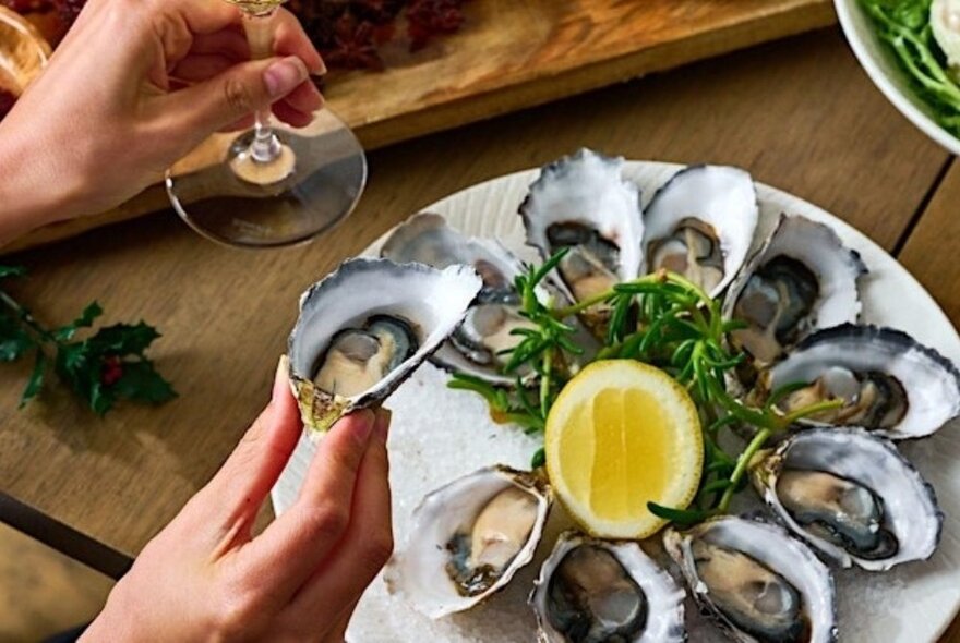 Hand holding a shucked oyster selected from a plate of freshly shucked oysters on a table, holding a glass in their other hand.