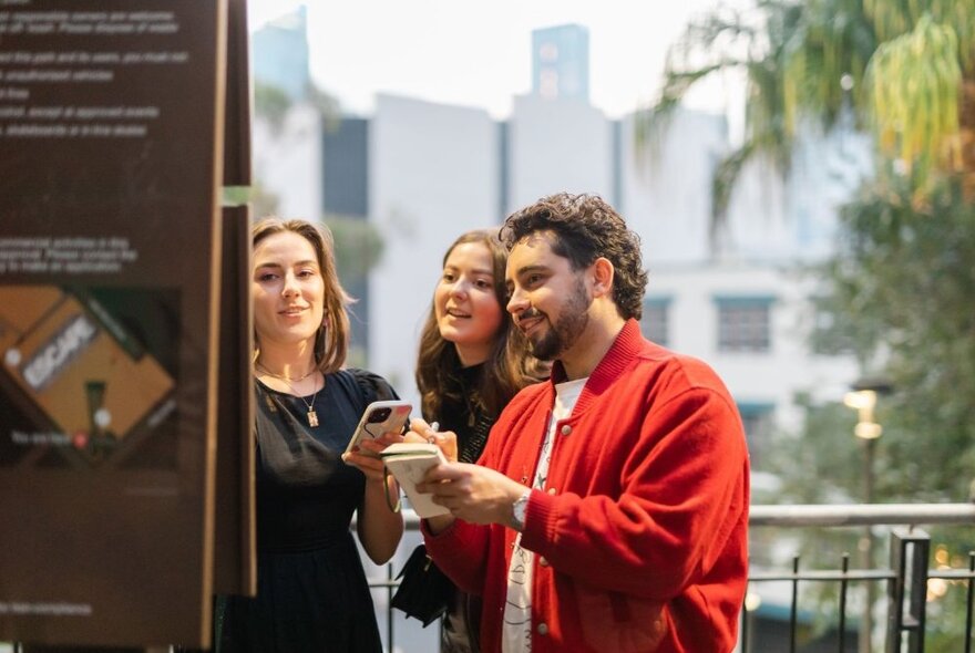 Three people standing in a city street, reading a sign, and one of them holding a smartphone. 