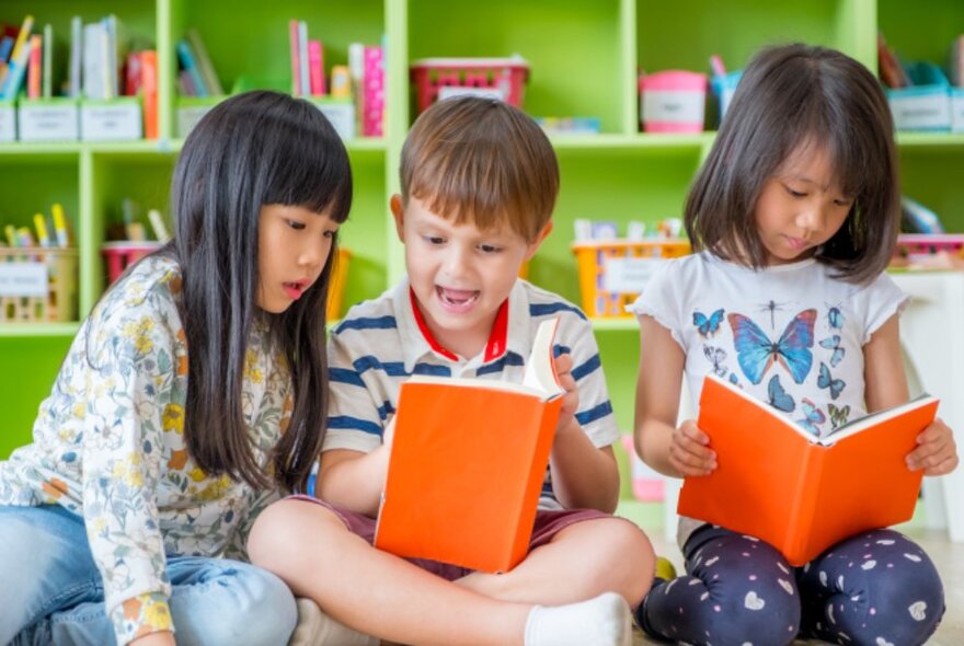 Three children sitting a library reading orange bound books. 