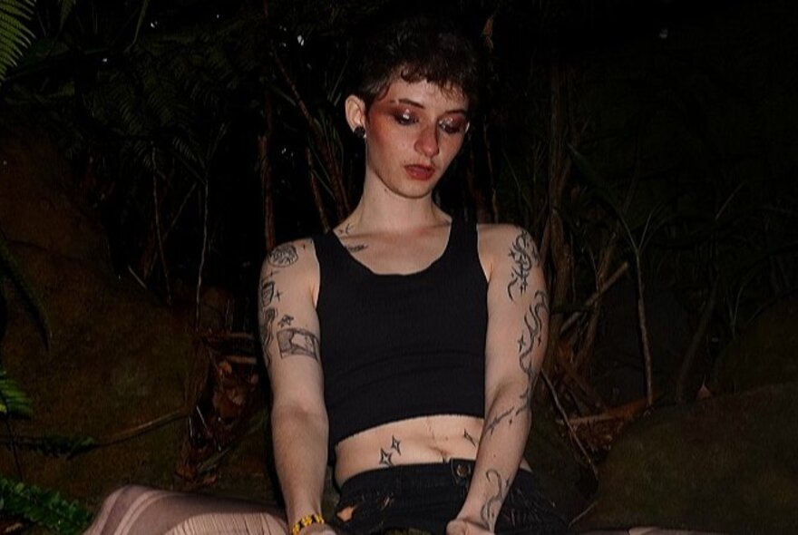 Boy with tattoos and short black top, looking downwards.
