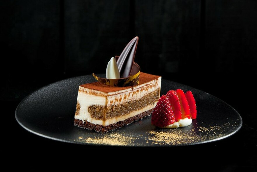 Chocolate oblong dessert on a black plate with strawberries to one side.