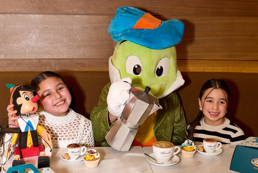 Two smiling children seated at a table in Brunetti's Carlton, with cupcakes and drinks, a Pinocchio wooden doll next to them and in the centre between the children person dressed in a green plushie costume pouring coffee into a cup.
