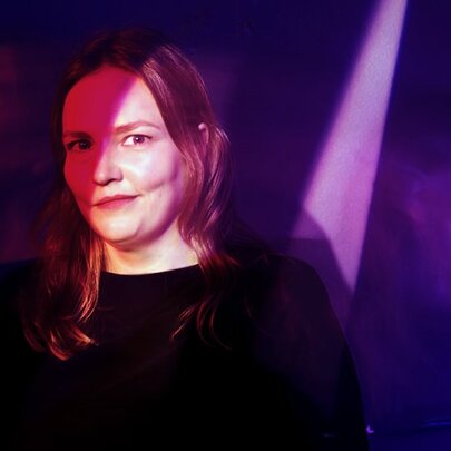 Comedian Kate Dehnert, with a wry smile, standing half in shadows, half in a spotlight, on a stage.