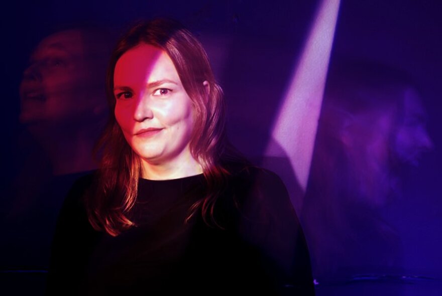 Comedian Kate Dehnert, with a wry smile, standing half in shadows, half in a spotlight, on a stage.