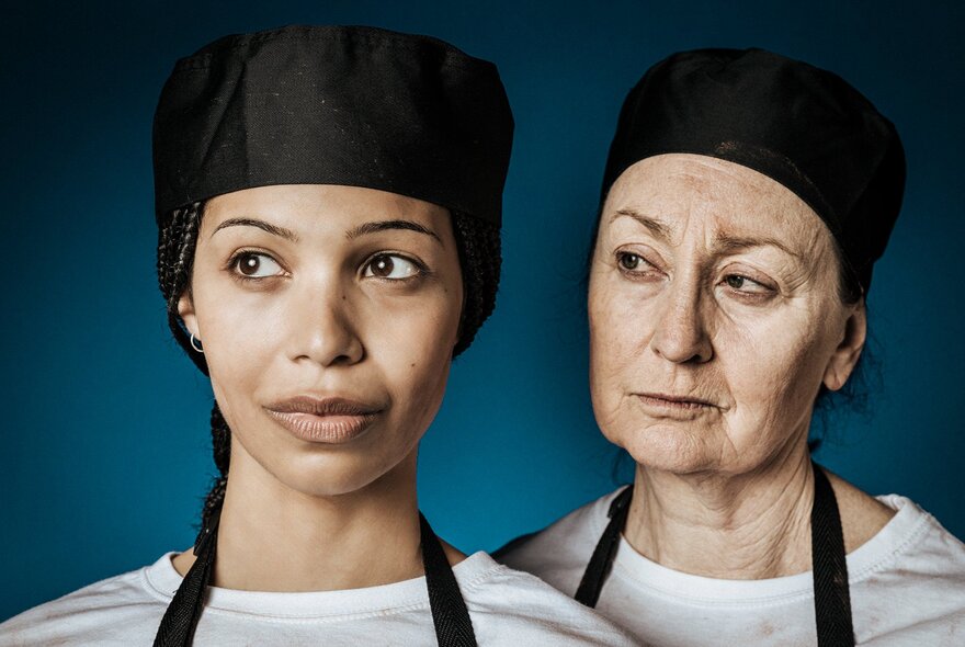 Two women wearing white t-shirts, black aprons and black deli caps, one younger and the other older.
