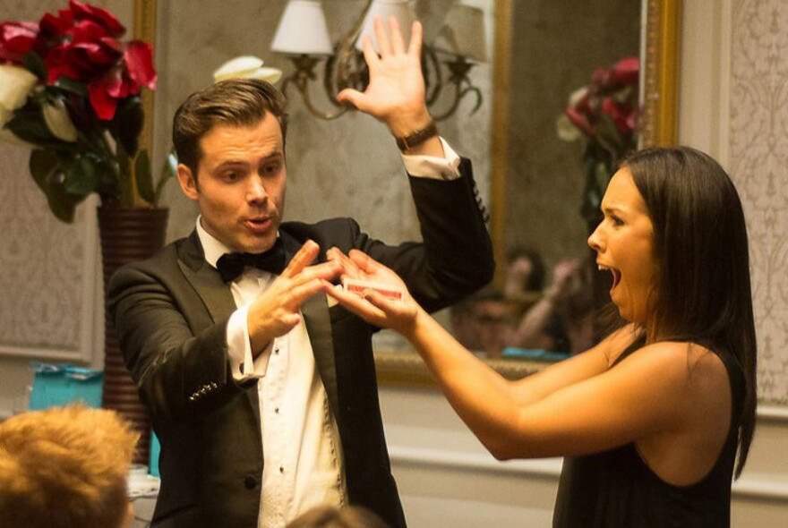 Man wearing a tuxedo performing a trick with an astonished woman, with lamps and mirrors in the background.